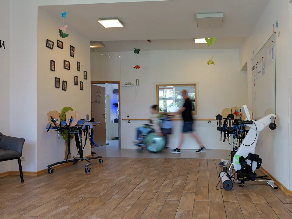 Kinder & Jugendliche bei Traumfänger. Ein großer geräumiger Flur im Inneren eines Traumfänger-Gebäudes. Eine Betreuungskraft schiebt eine Person im Rollstuhl. Es hängen Bilder in scharzen Rahmen an der Wand, gebastelte bunte Schmetterlinge hängen von der Decke