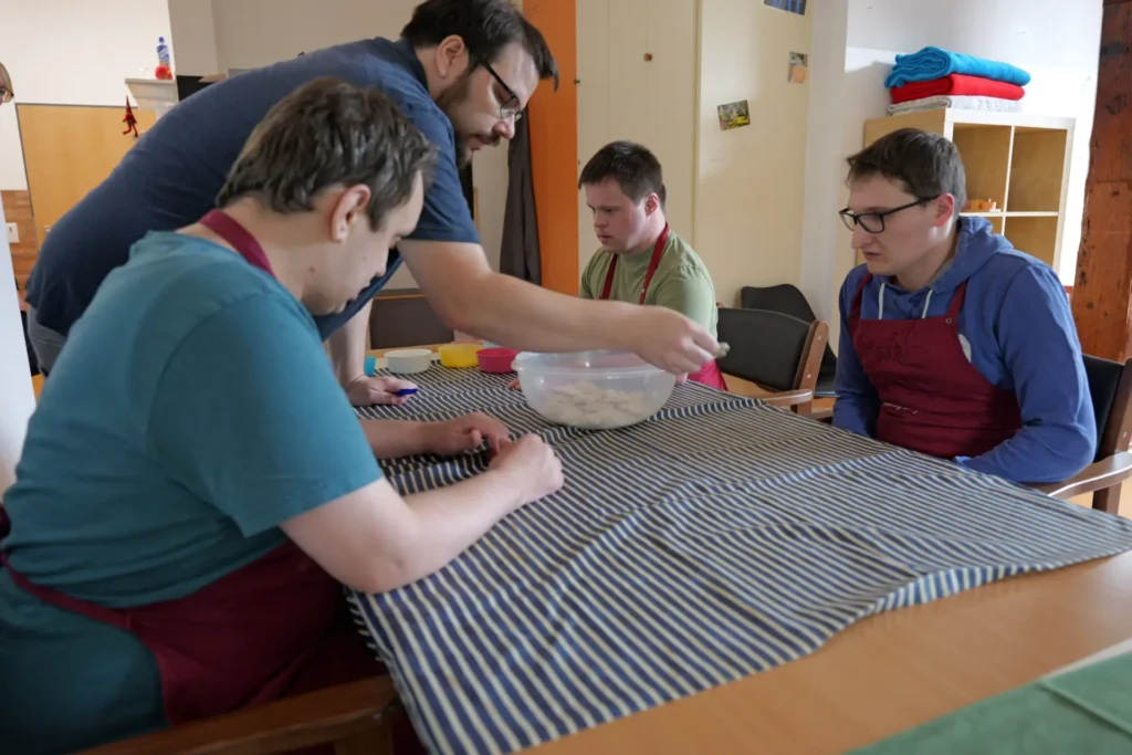 traumfaenger_teaser_team-werden Wohnen bei den Traumfängern . Drei erwachsene Männer sitzen mit Kochschürzen an einem Tisch. Eine stehende Betreuungskraft stellt eine Schüssel mit Keksen auf den Tisch